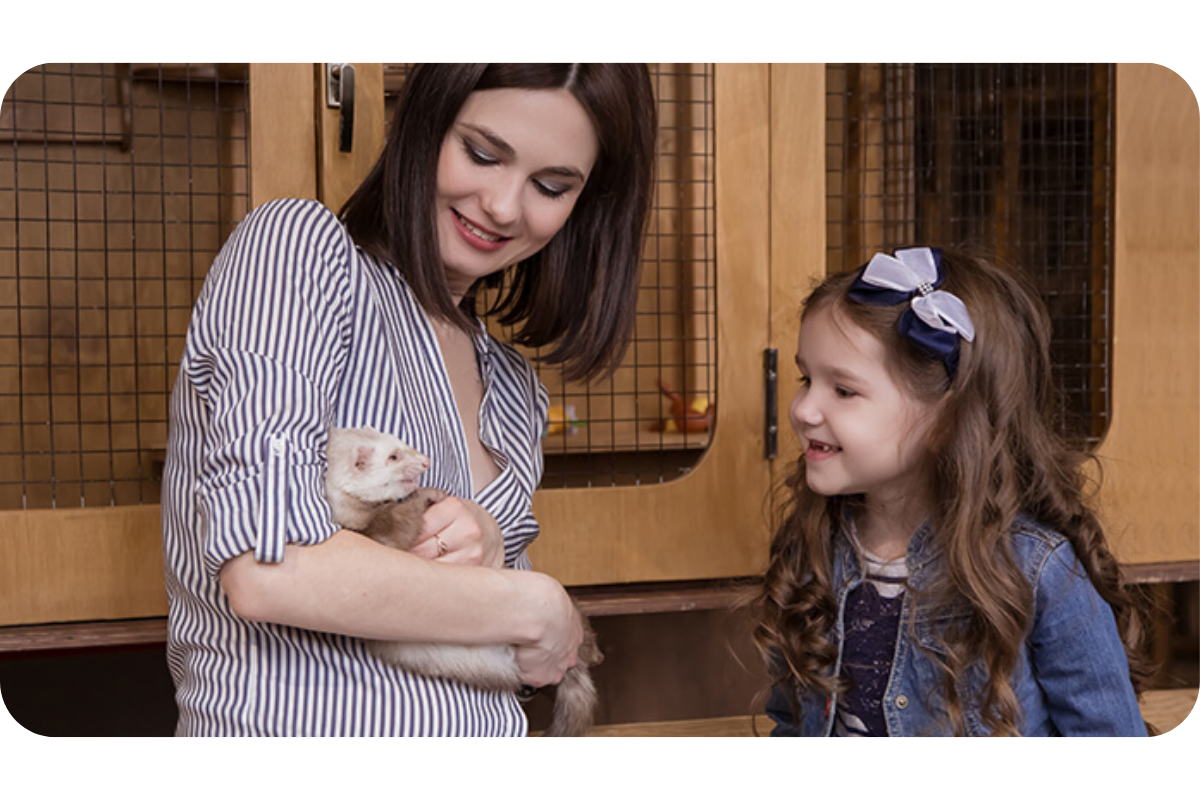 A woman cradles a ferret in her arms and shows it to a little girl beside her.
